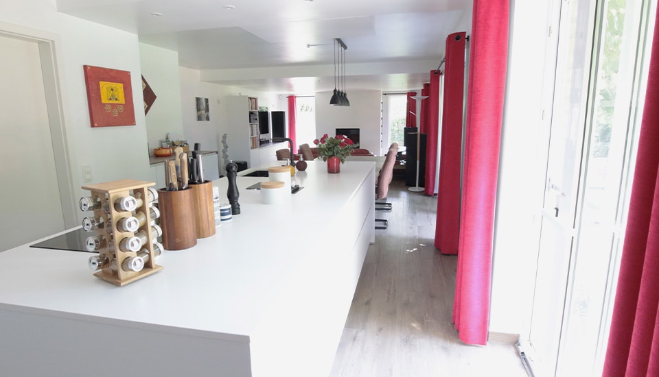 Cuisine moderne avec un îlot central blanc, équipé de pots à épices et accessoires de cuisine, entouré de rideaux rouges et baignant dans la lumière naturelle.