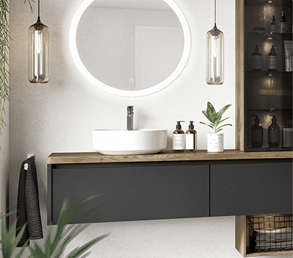 Un lavabo blanc sur un meuble noir avec un miroir rond éclairé, deux suspensions en verre et des plantes vertes.