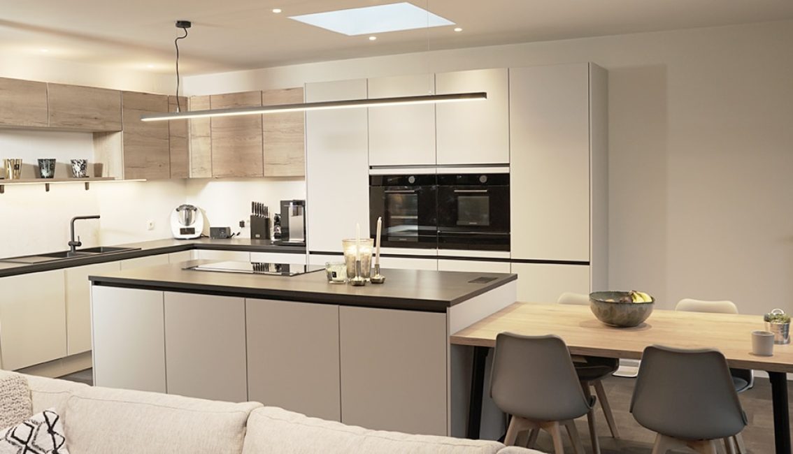 Cuisine moderne avec îlot central gris, meubles blancs et bois clair, équipée de fours encastrés, plan de travail noir et table à manger adjacente.